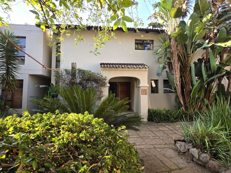 Front elevation of a townhouse featuring a covered entrance, stucco facade, and a stone paved walkway surrounded by established garden landscaping.