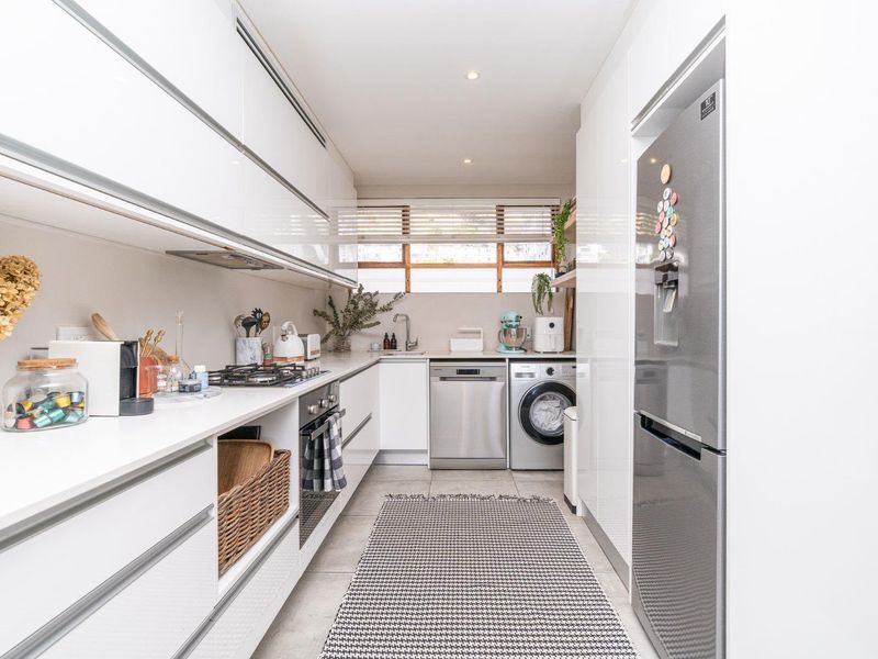 A modern, galley-style kitchen featuring sleek white cabinetry, integrated appliances, a gas hob, and tiled flooring with ample storage space.