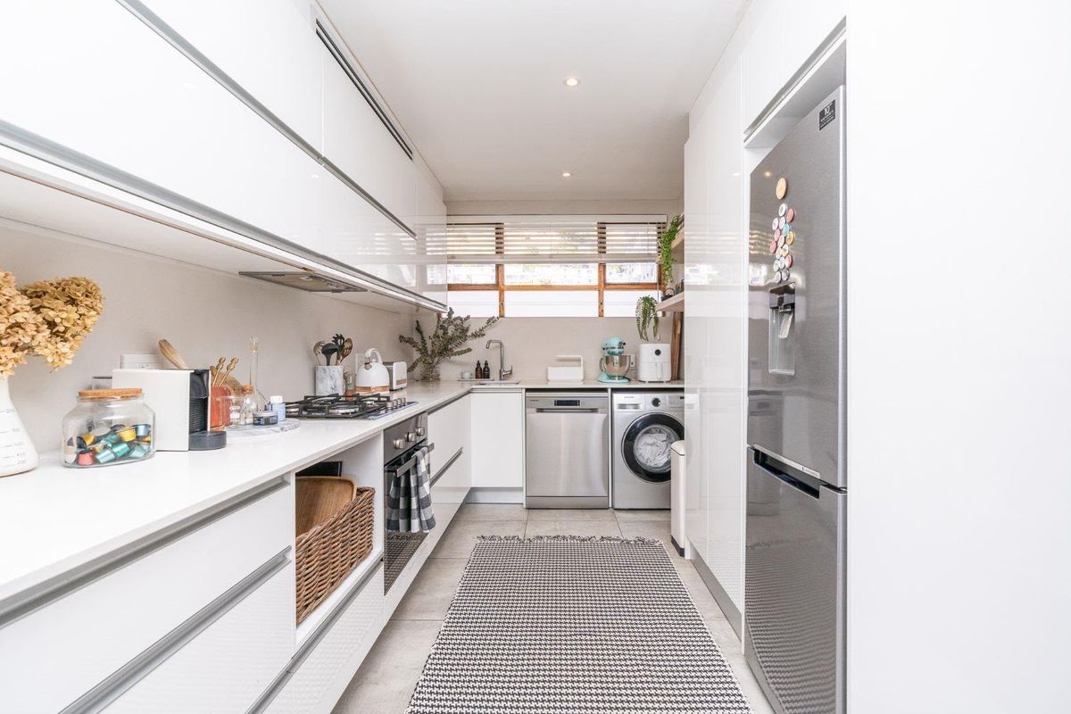 A modern, galley-style kitchen featuring sleek white cabinetry, integrated appliances, a gas hob, and tiled flooring with ample storage space.