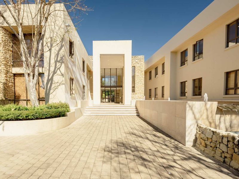 A modern office building entrance featuring a paved courtyard, stone-clad architectural details, and a central glass-fronted lobby with a water feature.