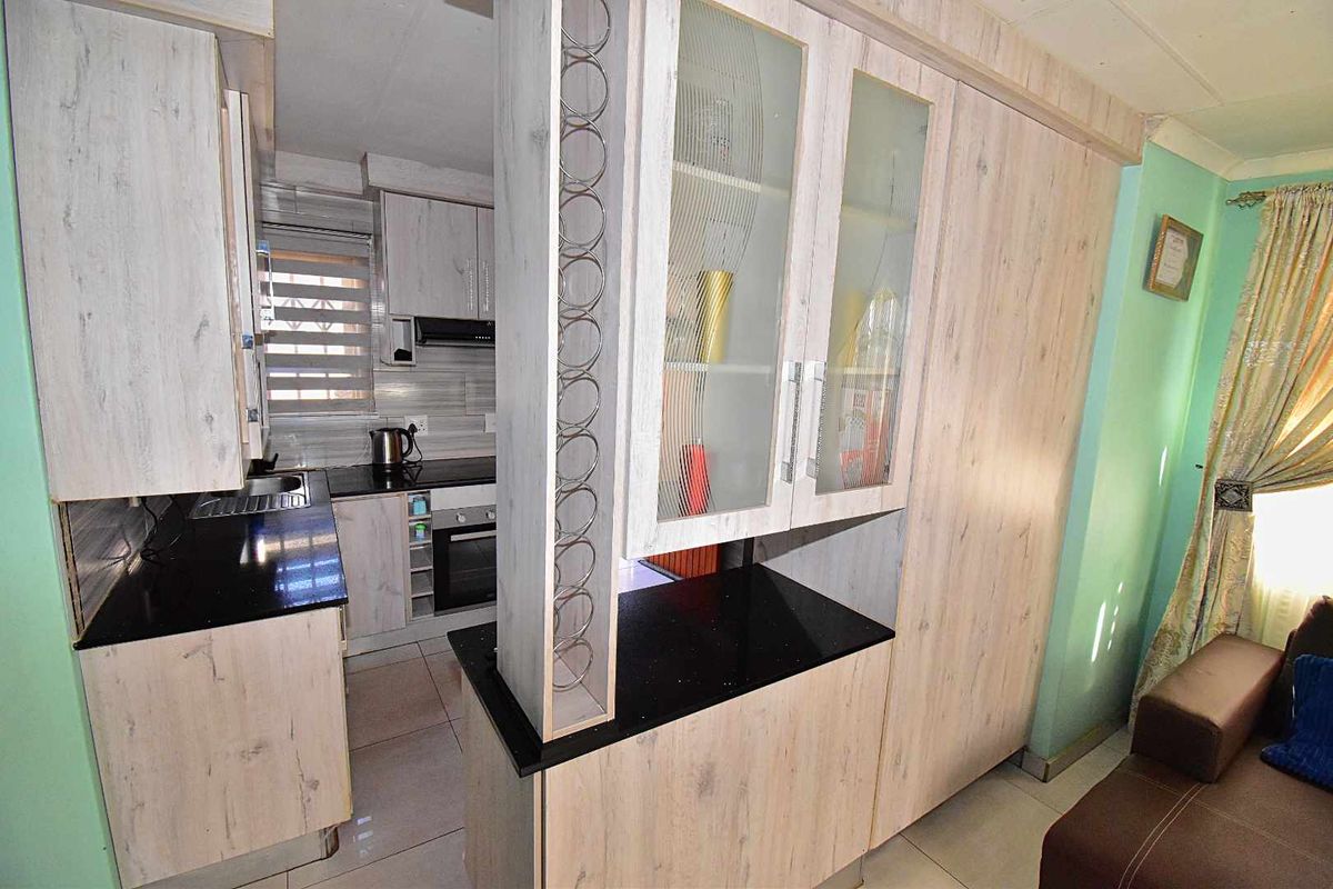 A compact kitchen featuring light wood-grain cabinetry, black countertops, a built-in oven, and a glass-fronted display cabinet with integrated wine rack.