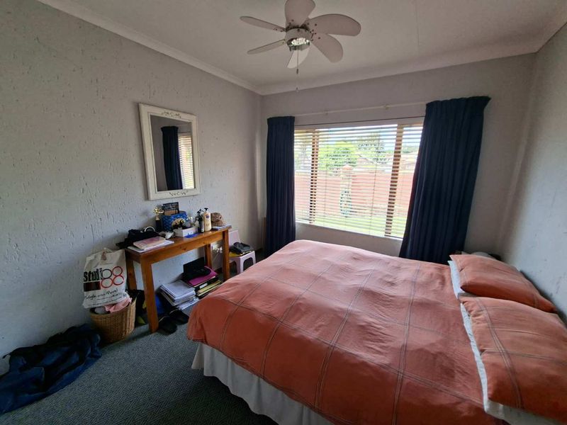 A standard bedroom featuring carpeted flooring, a ceiling fan, and large windows with venetian blinds, offering a functional space with potential for cosmetic updates.