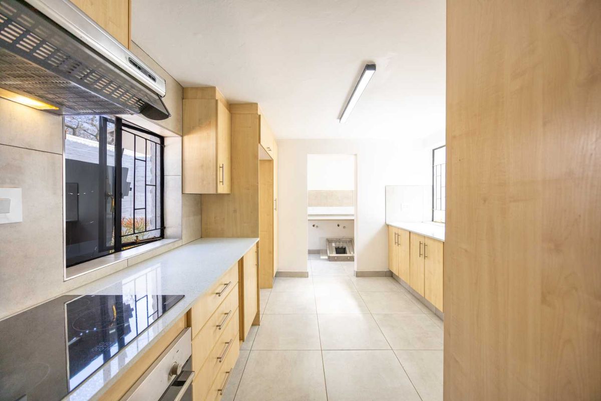 A clean, modern kitchen featuring light wood cabinetry, stone countertops, integrated appliances, and tiled flooring, leading into a secondary utility or scullery area.