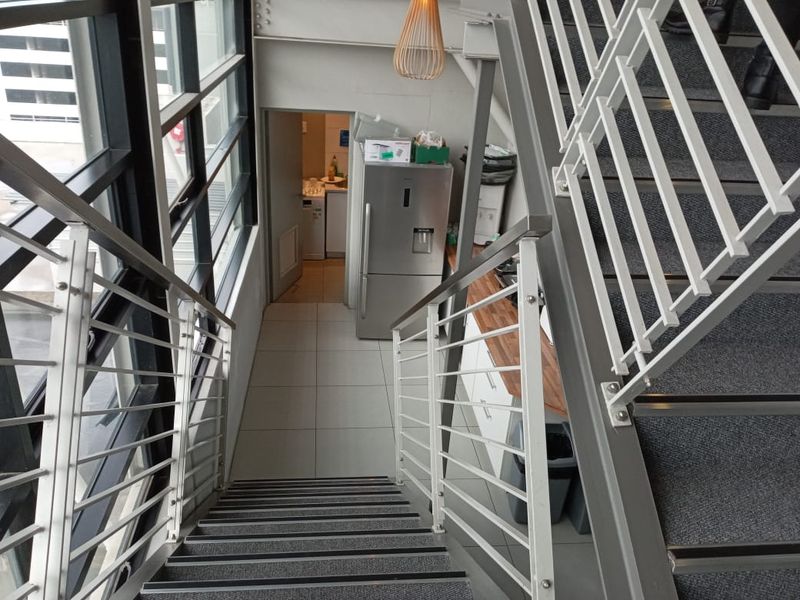 A modern office hallway featuring a metal staircase with balustrades, large glass windows, tiled flooring, and a compact kitchenette area with wooden countertops.
