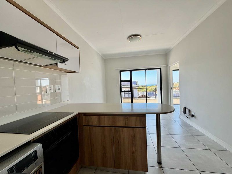 An open-plan kitchen and living area featuring modern cabinetry, built-in appliances, tiled floors, and a sliding door leading to a balcony with coastal views.