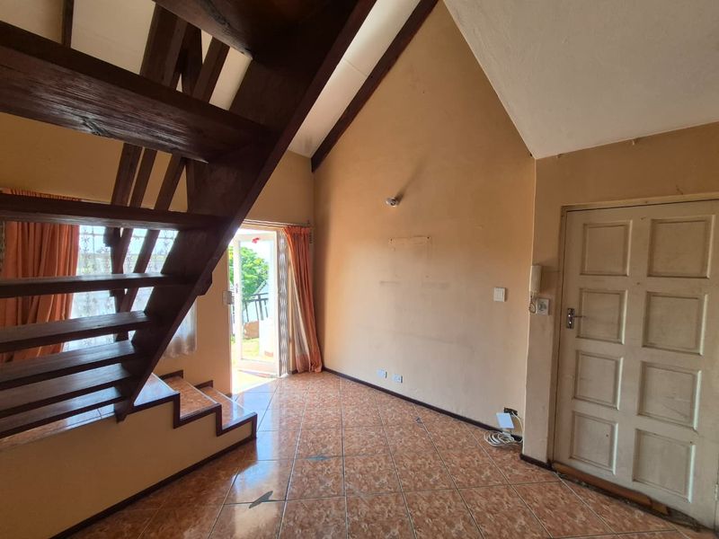 An open-plan living area featuring a prominent wooden staircase, vaulted ceilings, and tiled flooring, leading out to an exterior patio area.