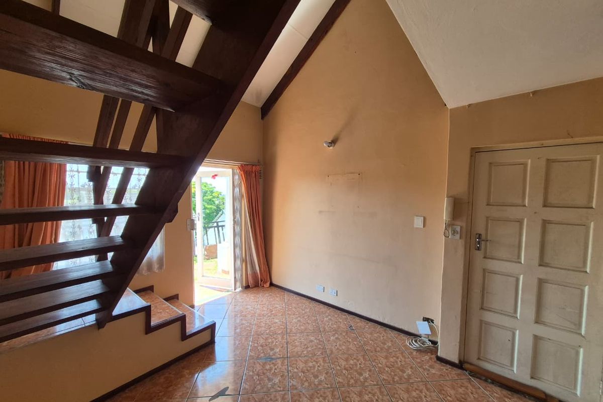 An open-plan living area featuring a prominent wooden staircase, vaulted ceilings, and tiled flooring, leading out to an exterior patio area.