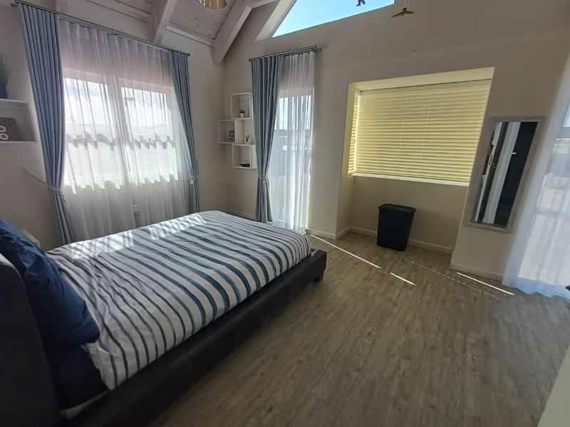 A bright bedroom featuring vaulted ceilings, exposed timber beams, wood-look flooring, and large windows with integrated blinds and curtains.
