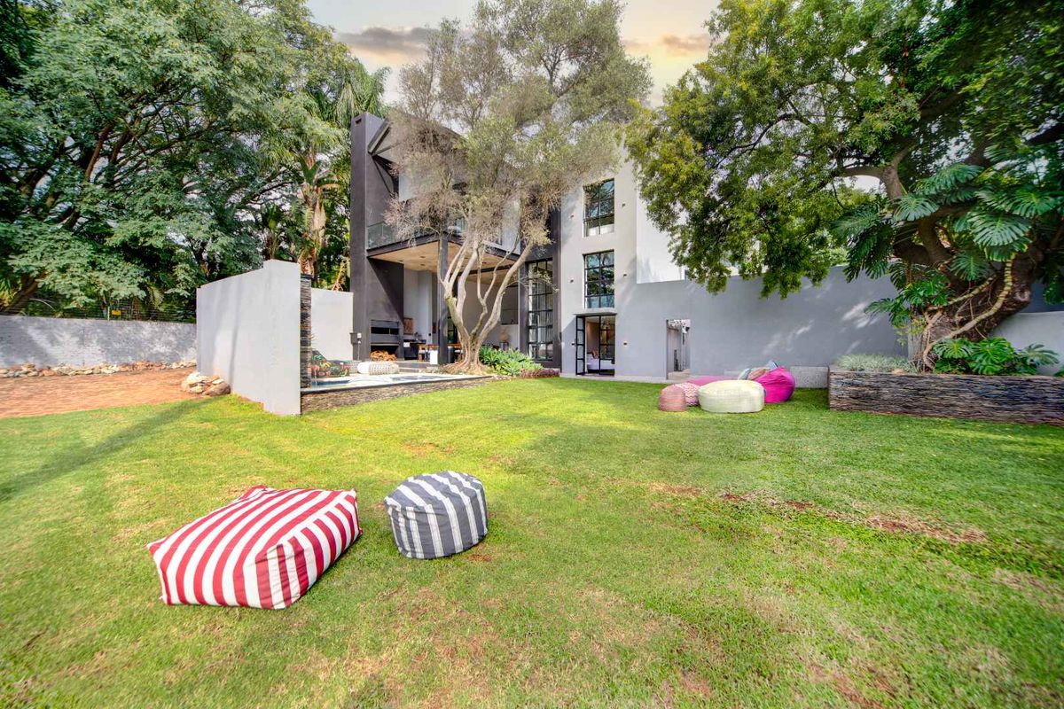 A modern multi-story rear facade featuring a covered patio, built-in braai area, and a well-maintained garden with mature trees.