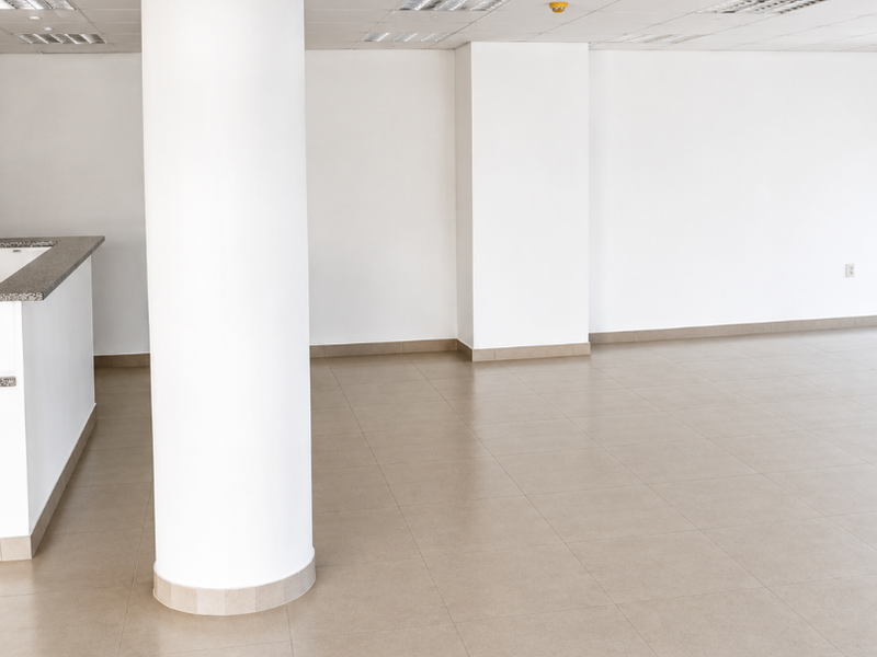 A spacious, empty office area featuring a neutral ceramic tile floor, a prominent structural column, and a built-in reception counter under a suspended ceiling.