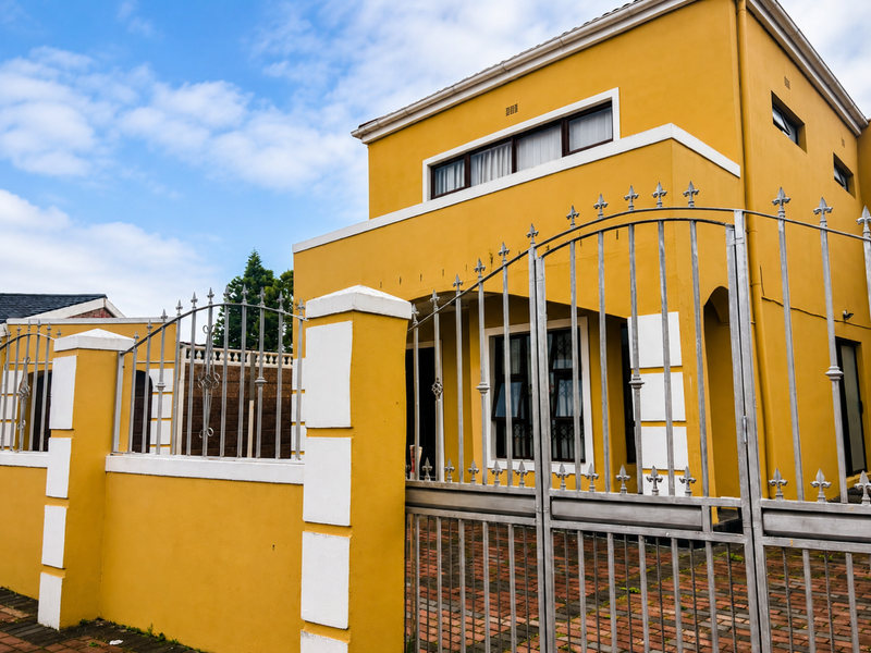 Front elevation of a two-story yellow house featuring a secure metal gate, masonry boundary wall, and paved driveway access.
