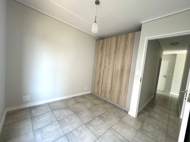 A clean, empty bedroom featuring modern wood-finish built-in cupboards, neutral grey tiled flooring, and a simple pendant light fixture.