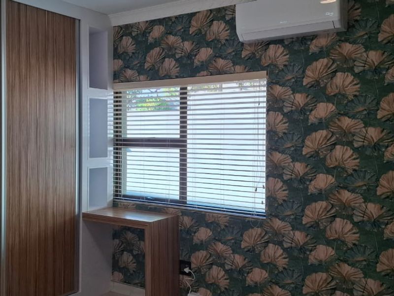 A well-maintained bedroom featuring built-in wooden wardrobes, a matching built-in desk with shelving, venetian blinds, and a wall-mounted air conditioning unit.