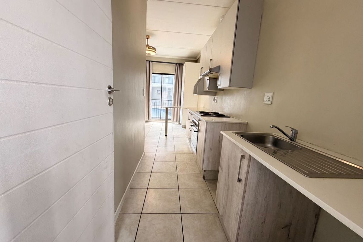 A functional galley-style kitchen featuring built-in cabinetry, a stainless steel sink, and integrated cooking appliances, leading towards a balcony area.