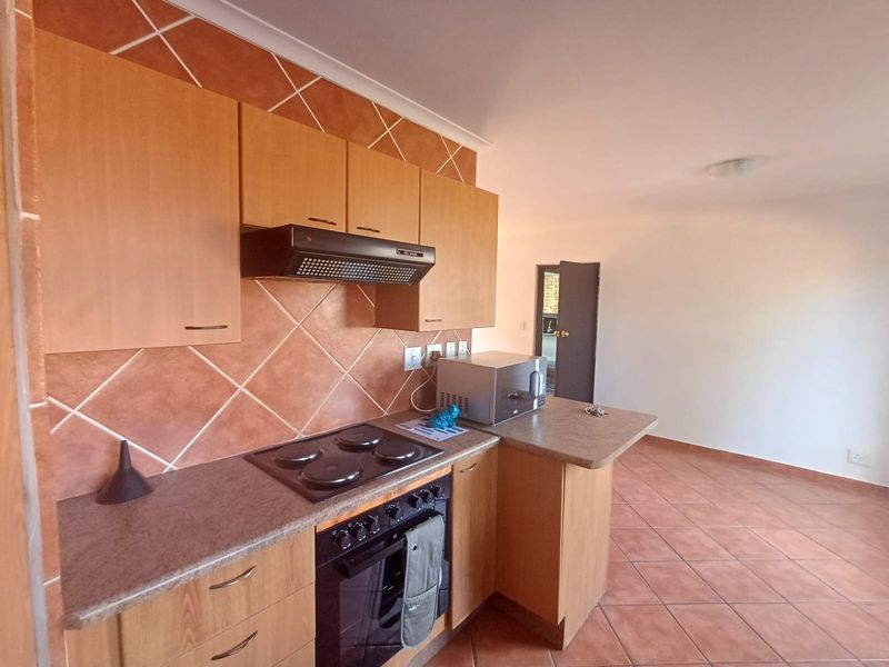 A functional kitchen featuring wood-finish cabinetry, tiled backsplash, and built-in cooking appliances, opening into an adjacent living area with tiled floors.