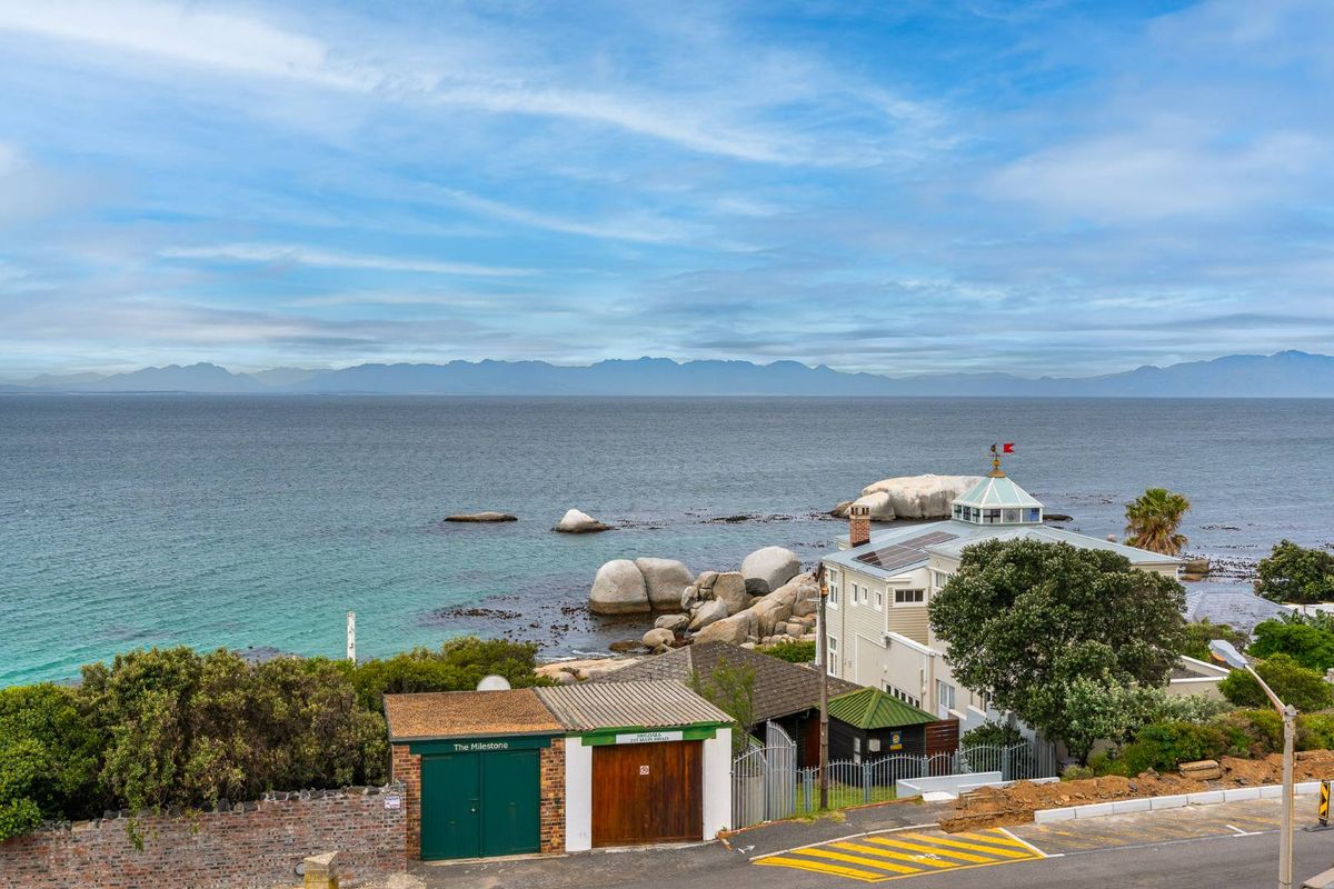 An elevated aerial view of a coastal property featuring a main residence with solar panels, garages, and a scenic ocean and mountain backdrop.
