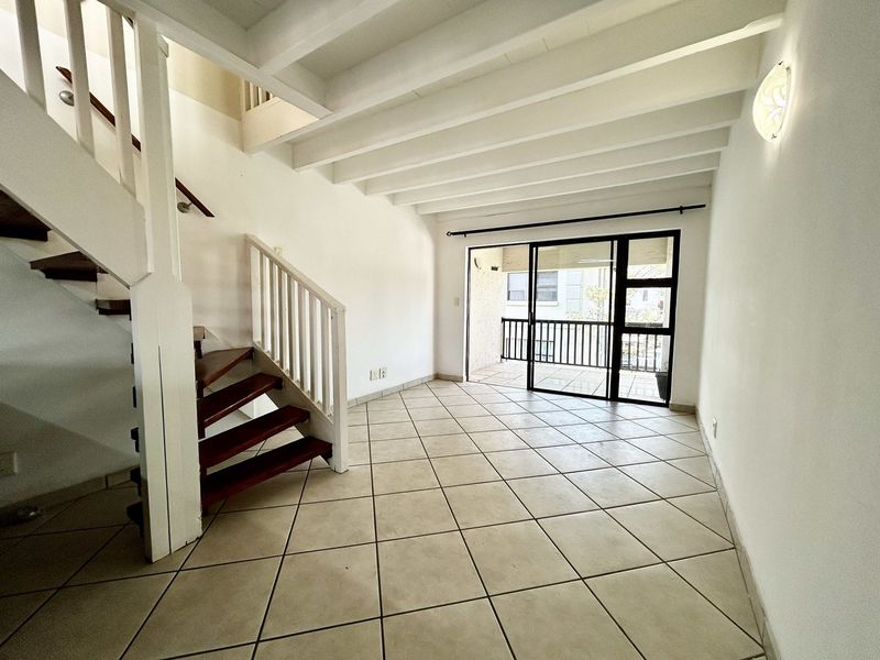 An empty, open-plan living area featuring a wooden staircase, exposed ceiling beams, tiled flooring, and direct access to a balcony through sliding glass doors.
