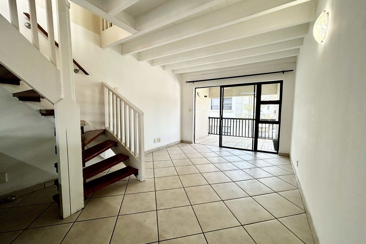 An empty, open-plan living area featuring a wooden staircase, exposed ceiling beams, tiled flooring, and direct access to a balcony through sliding glass doors.