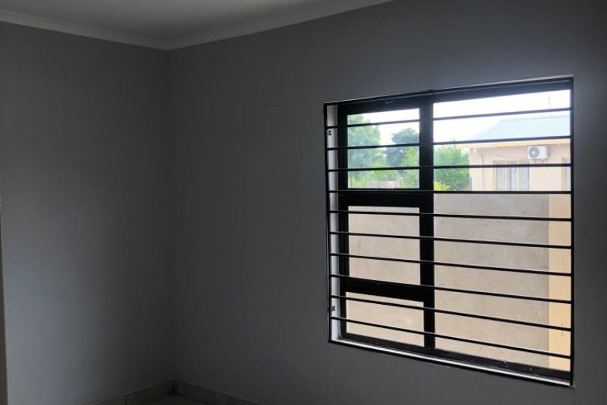 A clean, empty bedroom featuring neutral grey walls, tiled flooring, and a window fitted with security bars.