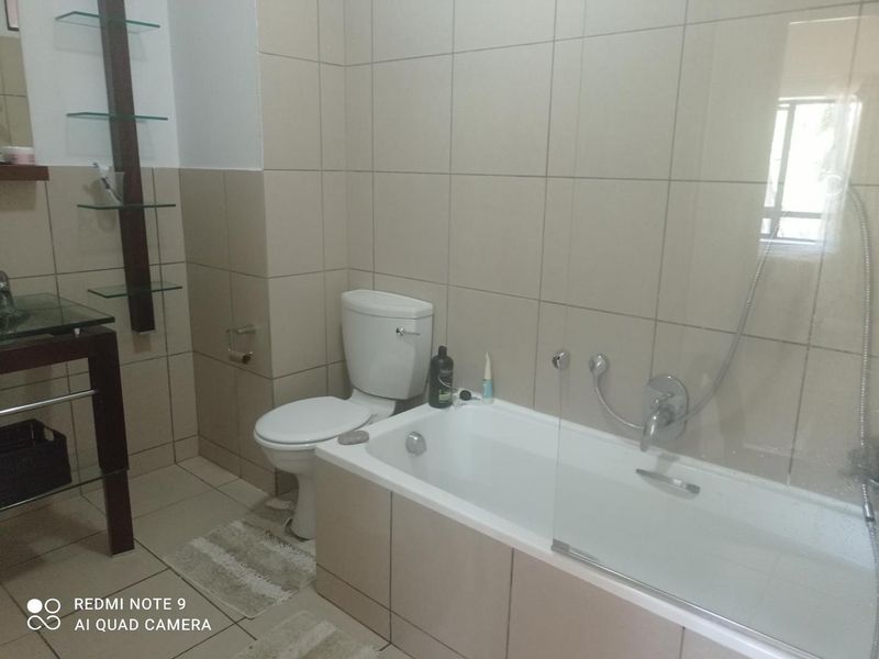 A functional bathroom featuring a bathtub with a shower mixer, glass screen, toilet, and wall-mounted shelving with neutral wall and floor tiling.