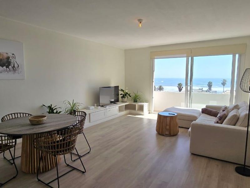 Bright open-plan living and dining area featuring light laminate flooring and large sliding doors leading to a balcony with ocean views.