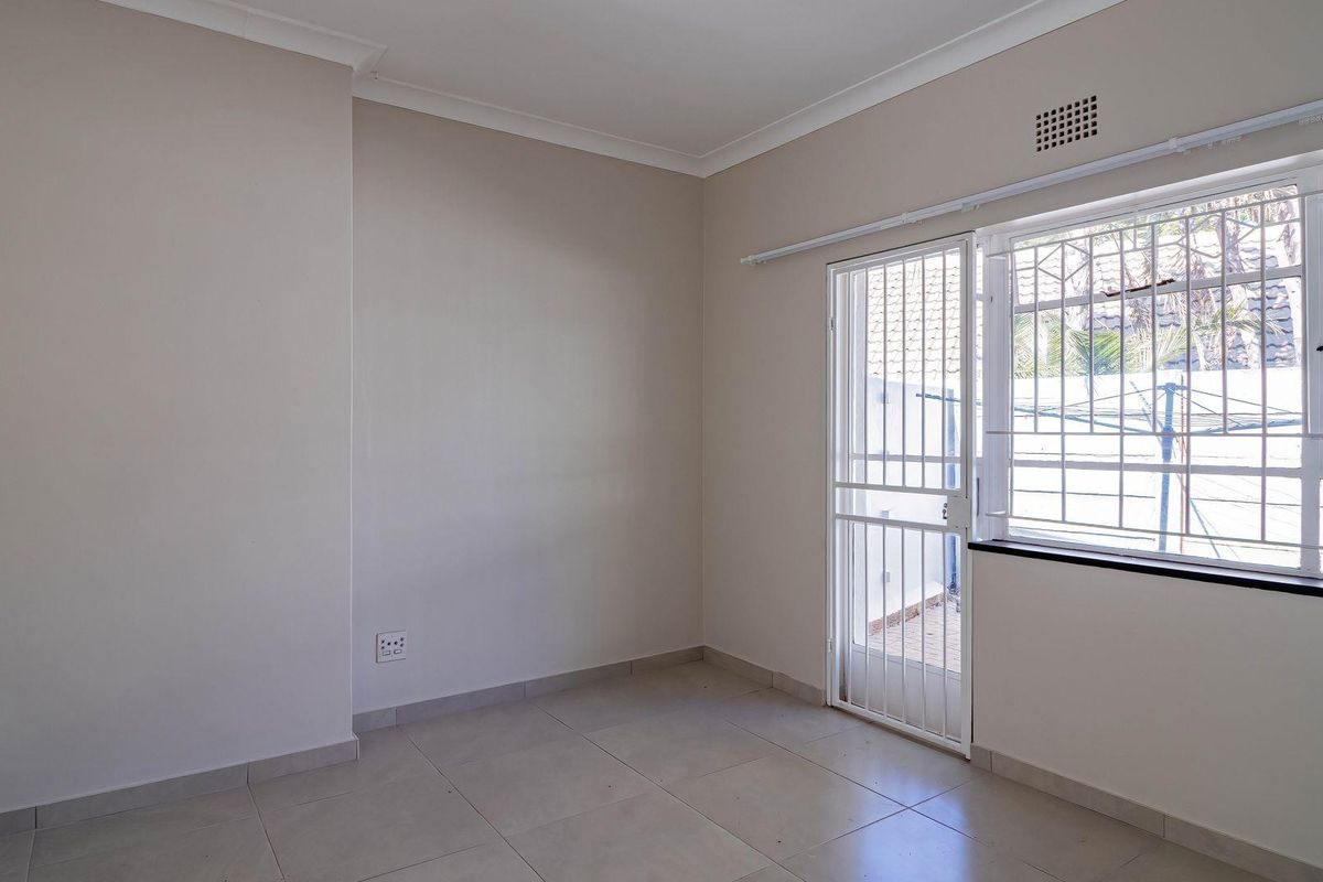 A bright, empty bedroom featuring neutral tiled flooring, a security gate leading to the exterior, and a window with security bars.