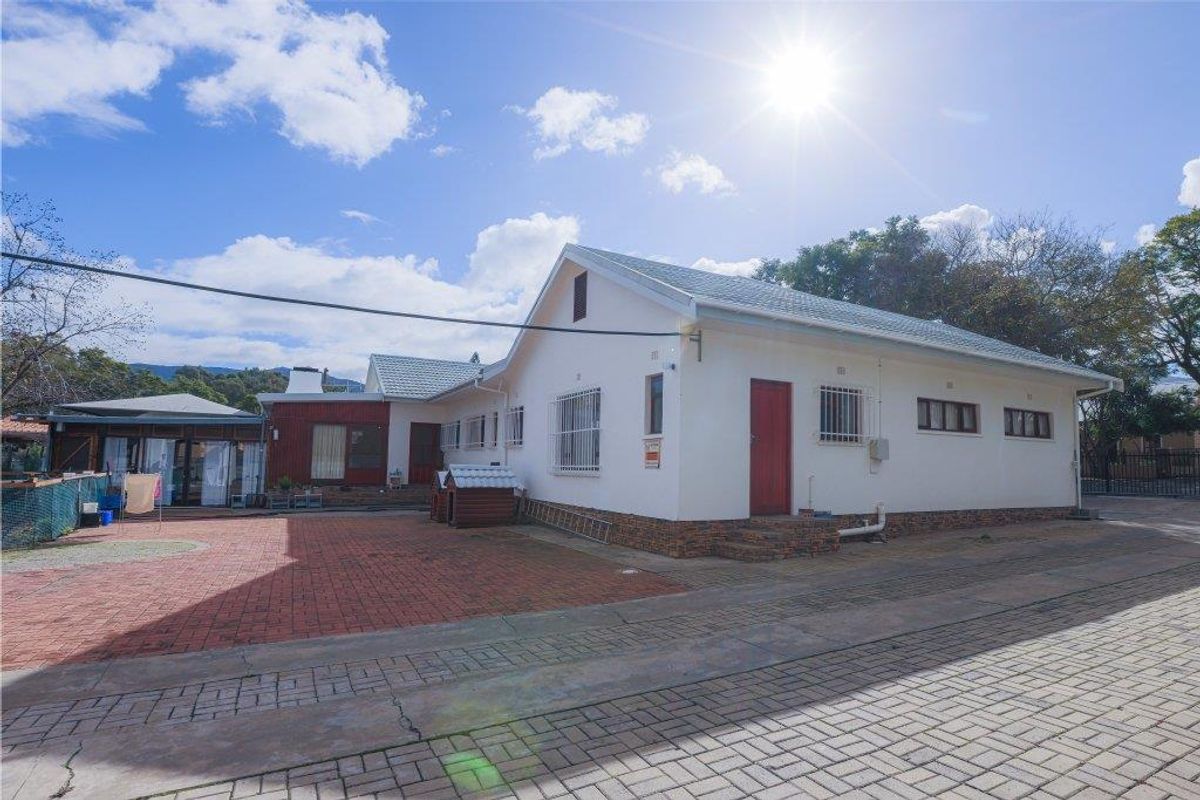Exterior view of the property featuring a paved courtyard, white facade, and red door, with a covered patio area in the background.