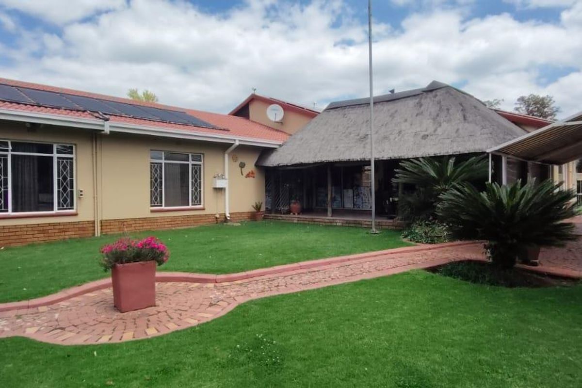 Rear view of the property featuring a lawn, paved walkway, carport, and a section with a traditional thatch roof and solar panels.