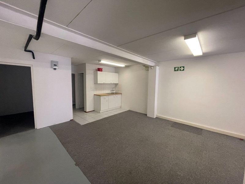A functional, open-plan office kitchenette featuring white cabinetry, a sink, and a mix of carpeted and tiled flooring in a commercial space.