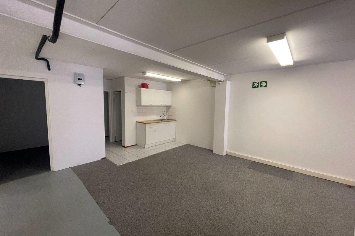 A functional, open-plan office kitchenette featuring white cabinetry, a sink, and a mix of carpeted and tiled flooring in a commercial space.