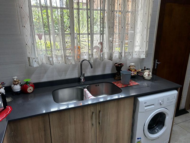 A functional kitchen area featuring a double stainless steel sink, dark stone countertops, wood-finish cabinetry, and a built-in space for a washing machine.