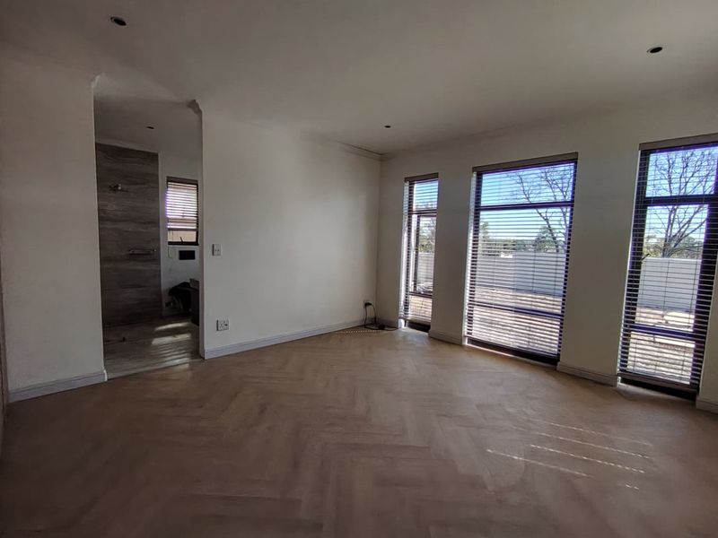 A bright, empty bedroom featuring modern herringbone flooring, recessed lighting, and large windows with venetian blinds, leading into an en-suite bathroom.