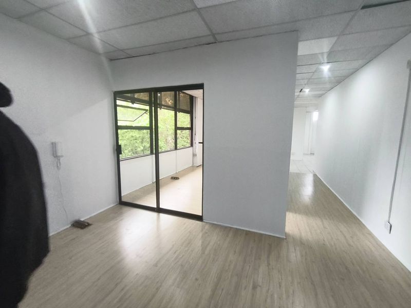 A bright, empty office space featuring light wood-look laminate flooring, a suspended ceiling, and a sliding glass door leading to an adjacent area.