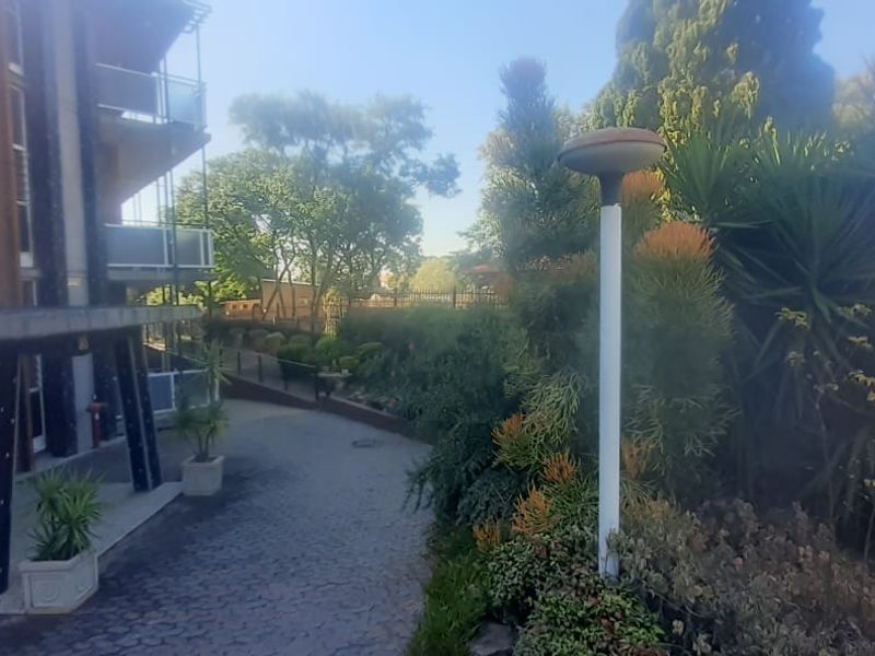 Exterior view of the apartment complex entrance featuring a paved driveway, landscaped garden beds, and secure metal perimeter fencing.