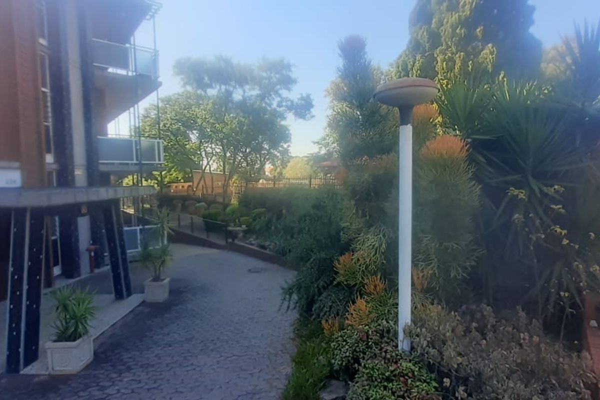 Exterior view of the apartment complex entrance featuring a paved driveway, landscaped garden beds, and secure metal perimeter fencing.
