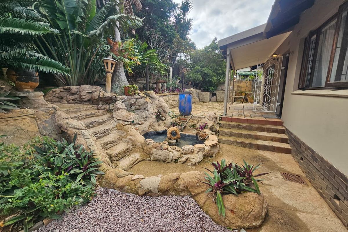 Rear exterior view featuring a landscaped garden with a stone-clad water feature, paved patio area, and concrete steps leading to the house entrance.