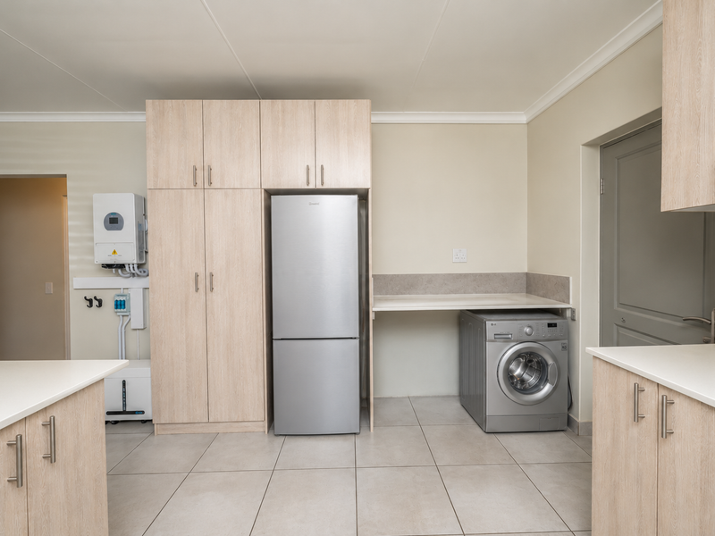 A functional scullery featuring light wood cabinetry, stone countertops, tiled flooring, and a dedicated wall-mounted inverter and battery system for power backup.