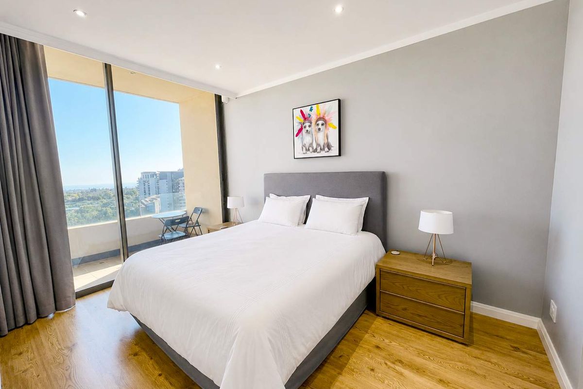 A bright and modern bedroom featuring laminate flooring, recessed downlights, and large sliding glass doors providing access to a private balcony with scenic views.