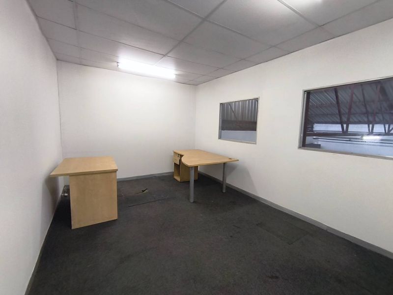 A functional, empty office space featuring a suspended ceiling, fluorescent lighting, and internal windows overlooking the industrial warehouse area.