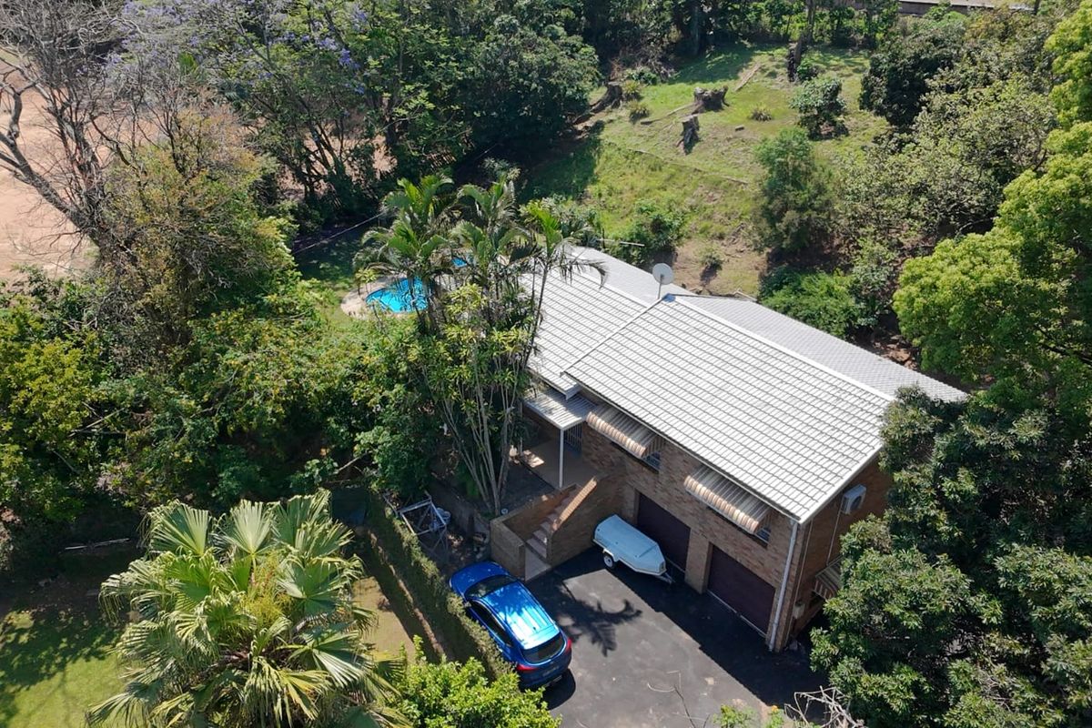 Aerial view of a residential property featuring a tiled roof, double garage, asphalt driveway, and a private swimming pool set within a lush garden.