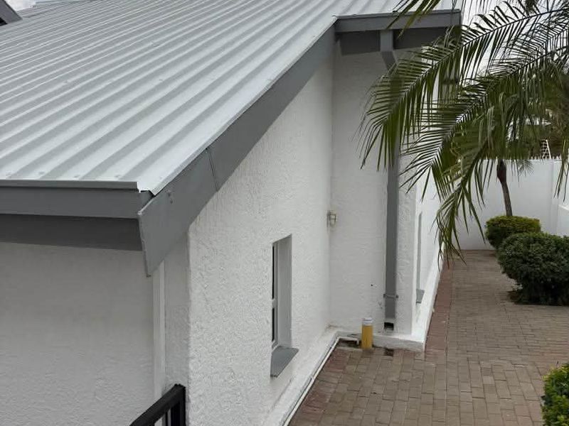 A view of the rear exterior featuring a corrugated metal roof, textured white walls, a paved walkway, and a metal railing.