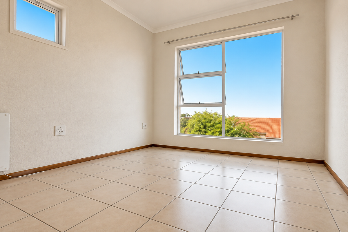 A bright, empty bedroom featuring neutral tiled flooring, large aluminium windows, and a wall-mounted electric heater, offering a clean and versatile living space.