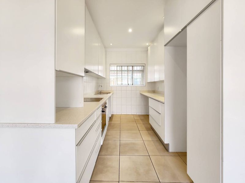 A clean, modern galley-style kitchen featuring white cabinetry, stone countertops, built-in oven, and tiled finishes throughout.