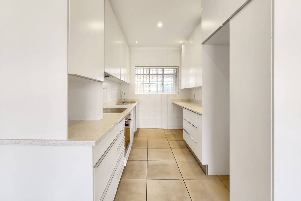 A clean, modern galley-style kitchen featuring white cabinetry, stone countertops, built-in oven, and tiled finishes throughout.