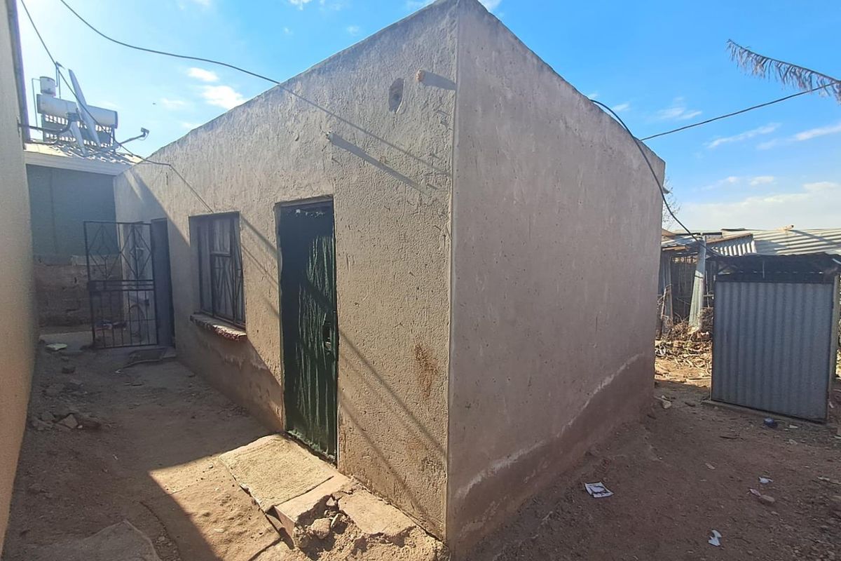 Rear exterior view of a basic residential structure featuring a simple concrete facade, security gate, and metal window frame.