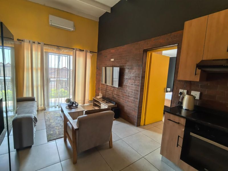 Open-plan living area featuring an exposed brick feature wall, tiled flooring, and a compact kitchen with built-in cabinetry and integrated appliances.