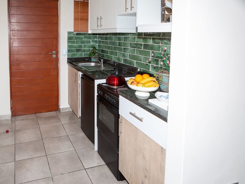 A compact kitchen featuring modern white and wood-tone cabinetry, dark granite countertops, a green tiled backsplash, and tiled flooring.
