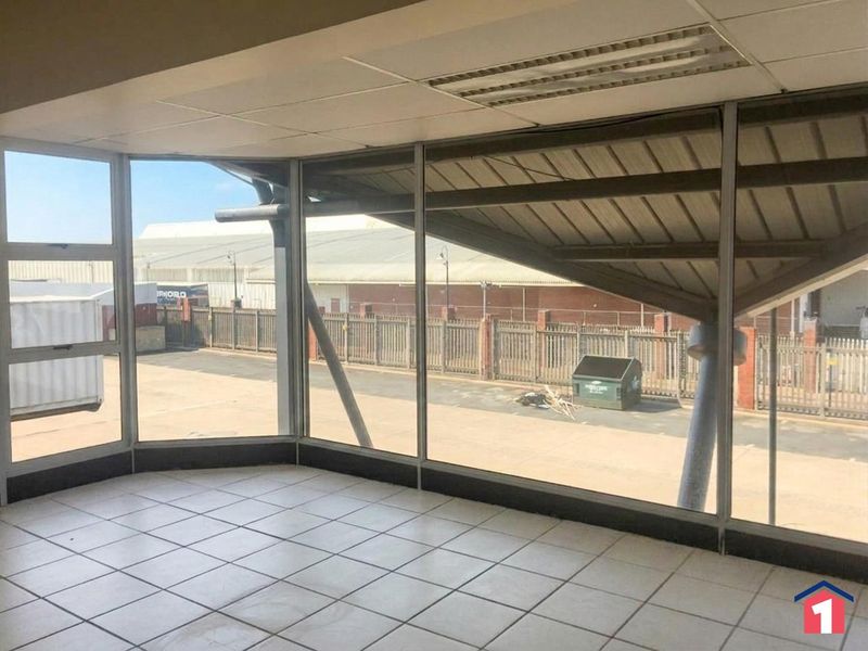 An empty office space featuring large glass windows, tiled flooring, and a suspended ceiling with recessed lighting, overlooking an industrial yard.
