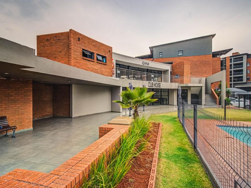 Exterior view of the complex clubhouse featuring a covered patio area, brick architecture, and access to the communal swimming pool and garden facilities.