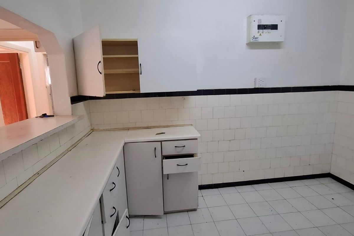 An empty, dated kitchen featuring white tiled walls, tiled flooring, and built-in cabinetry with a convenient pass-through window to the adjacent living area.